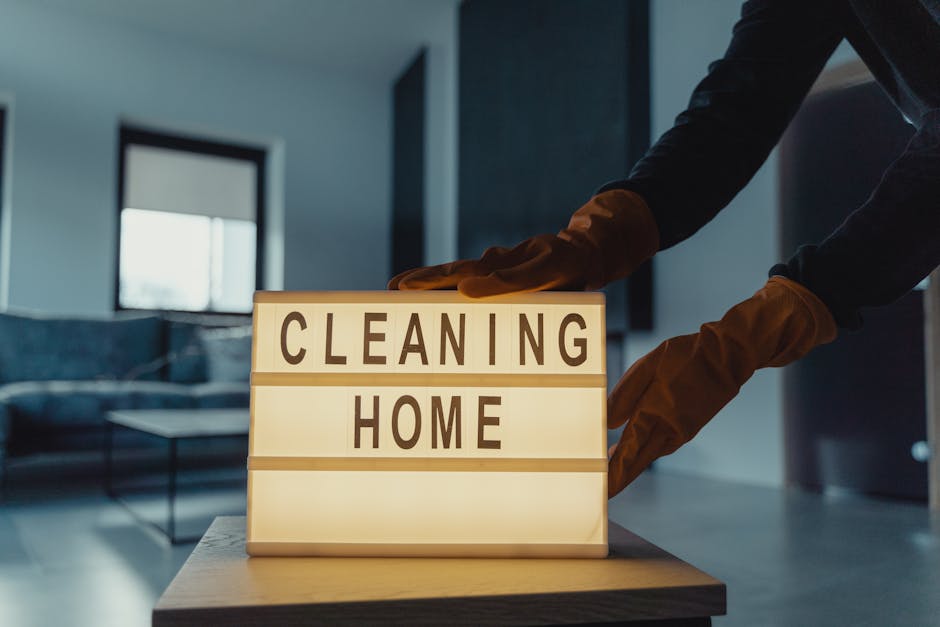 An orange plastic bucket placed on a smooth, grey floor next to a wall, with a blue microfiber cleaning cloth draped over the edge of the bucket. A mop with a blue handle and a flat cleaning head is l