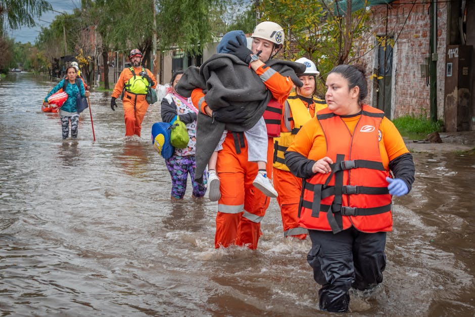 Rescue workers in orange waterproof suits and safety helmets assisting residents through floodwaters in an urban street, with water reaching above their ankles. In the background, additional rescue pe