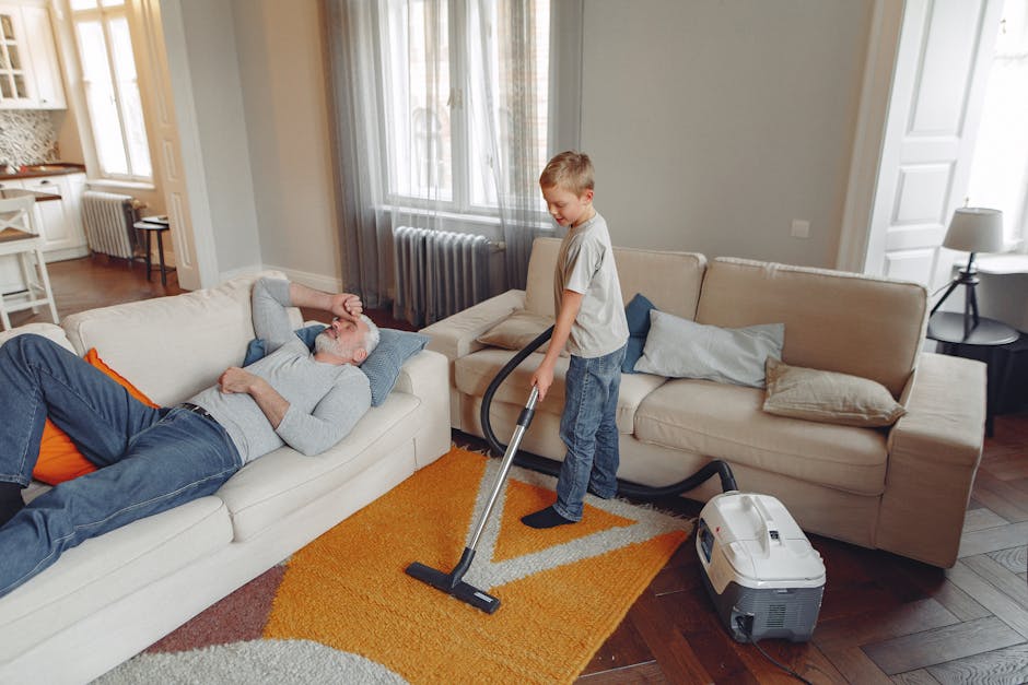 A person wearing full protective gear, including a white disposable suit, face mask, gloves, and shoe covers, is conducting deep cleaning on a beige, textured rug in a living room. The individual is u