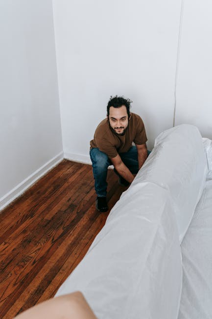 A man with curly dark hair and a beard, dressed in an orange sweater and grey pants, is kneeling on a wooden floor in a well-lit room, carefully cleaning a textured beige carpet with his right hand. T